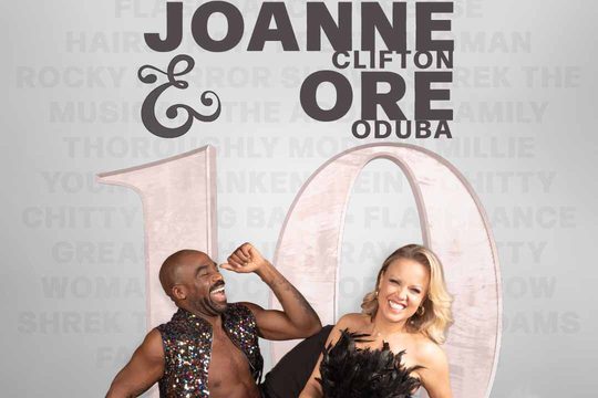 A black man in a sparkly jacket and a white, blonde woman in a black dress with feathers. They are laughing.