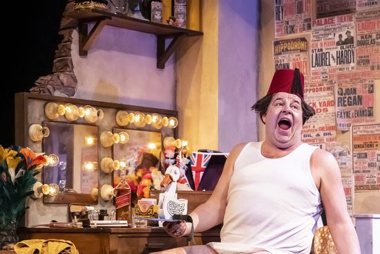 A man in a white vest and a Fez sitting down and laughing with his mouth open.