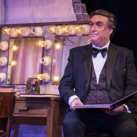A man in a suit, waistcoat and bow tie sits reading a book in front of a dressing room mirror.