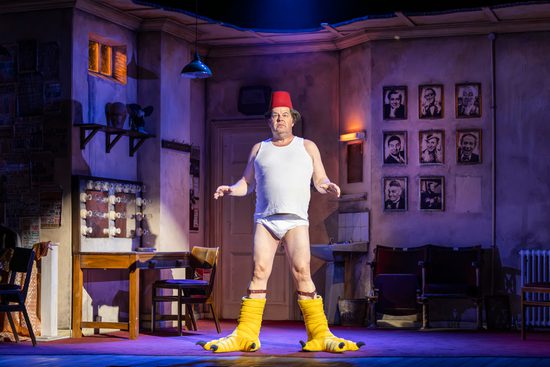 An older white man stands in the centre of a set made to look like a dressing room. He wears a white vest and underwear, large bird-like feet and a fez.