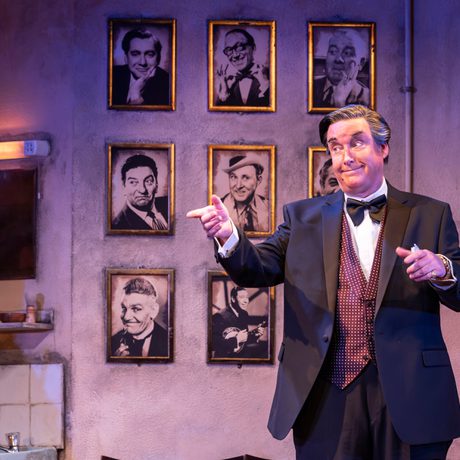 A man in a black suit and burgundy waistcoat points to the side. Behind him on the wall are black and white headshots in gold frames.
