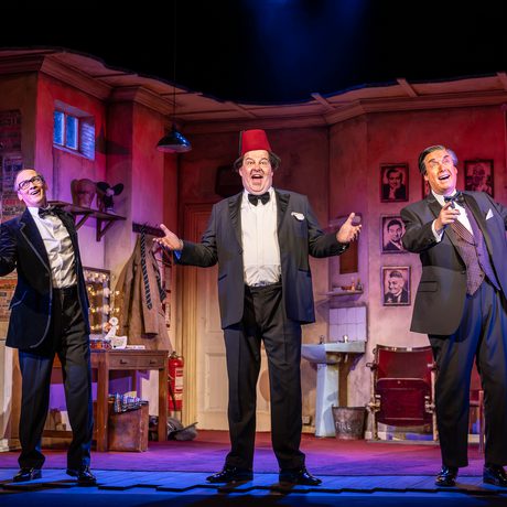 Three older white men in a dressing room, looking out towards the audience and smiling. The man in the centre wears a fez.