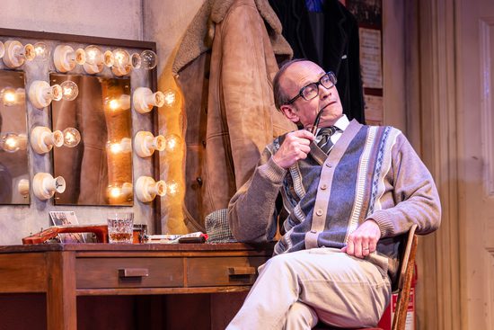 An older white man with black glasses, wearing a beige knitted cardigan. He is sat in front of a dressing room mirror with a pipe in his mouth.