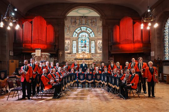 A brass band wearing red jackets and holding their instruments. They are standing in a church. The organ pipes in the background are lit red.