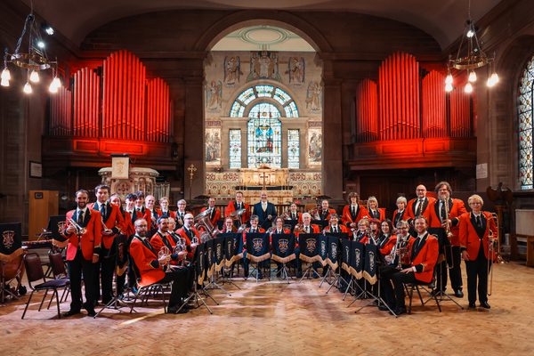 A brass band wearing red jackets and holding their instruments. They are standing in a church. The organ pipes in the background are lit red.