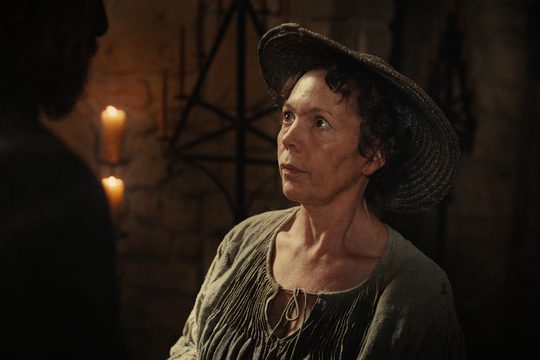 A woman with short brown hair wears a grey dress and a wide brimmed hat. She is standing in a candlelit room.