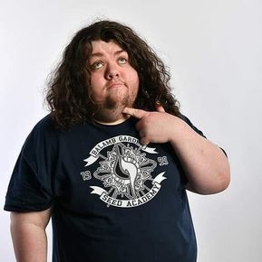 A white man with long, brown curly hair wearing a black t-shirt holds a finger to his chin, as if he is thinking.