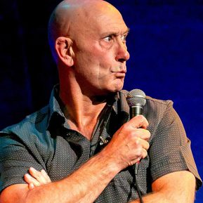 A bald, white man holds a microphone and pouts his lips, looking to the side.