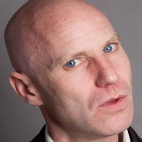 A bald, white man looks straight at the camera.