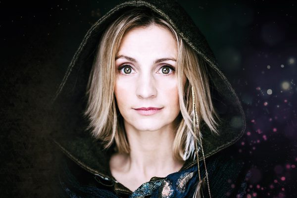 White woman with blonde hair and green eyes wears a black hood and looks to camera