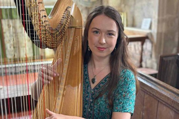 A woman with long brown hair wearing a green dress. She is holding a large harp.