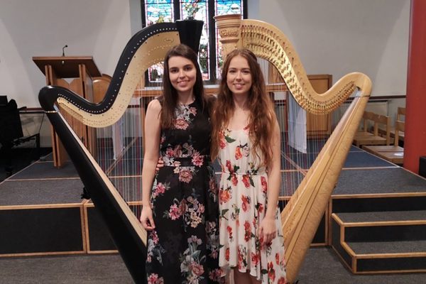 Two women in flowery dresses standing in front of two harps.