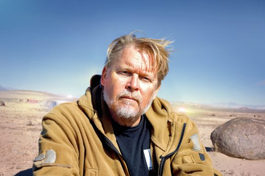 Man with blond hair and grey bears looks to the camara. Behind him a desert landscape