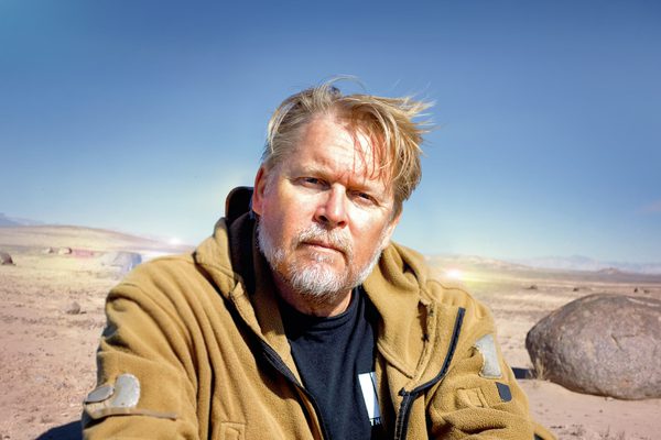 Man with blond hair and grey bears looks to the camara. Behind him a desert landscape