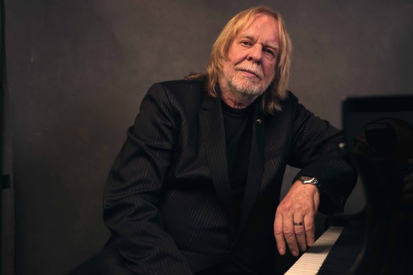 An older man with shoulder length blonde hair and a beard. He is wearing a black suit and sitting at a piano.