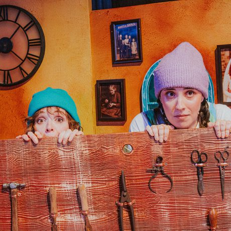 Two actors in colourful woollen hats look scared and peek over a wooden workbench covered in tools.