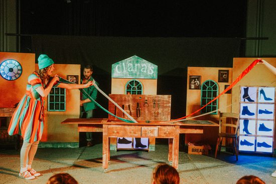 Actors in brightly coloured clothing pull long colourful ribbons from a wooden table.
