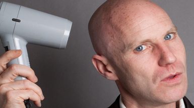 A bald man holding a hairdryer to his head.