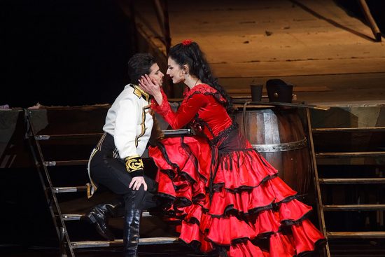 A man in a white and gold top sitting on stairs with a lady in a red dress, who is holding his face.