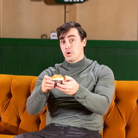 Man holding muffin up to camera sat on an orange sofa