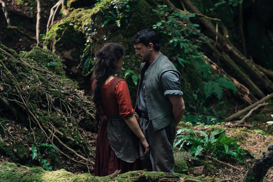 A man wearing blue and a woman wearing red stand close to each other in the woods.