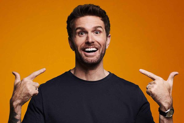 Joel Dommett in a black t-shirt, pointing to his face with both hands and smiling. He is in front of a bright orange background.