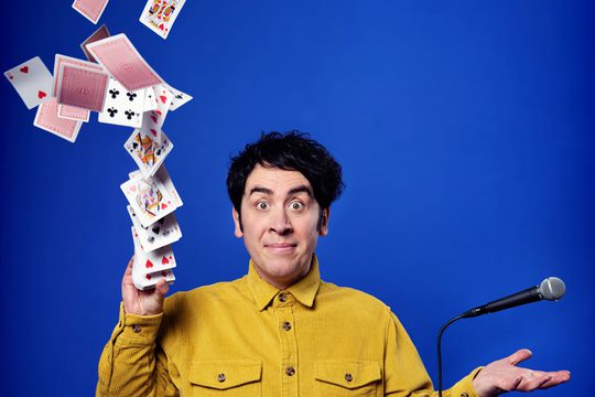 Pete Firman in a yellow shirt, with a microphone in one hand and a deck of cards coming from the other.