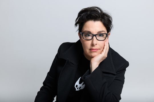 Sue Perkins wearing black glasses and a black coat, sitting with her chin resting on one hand.