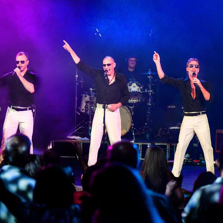 Three men in black shirts and white trousers. They are all holding a microphone in one hand, with the other hand pointing in the air.