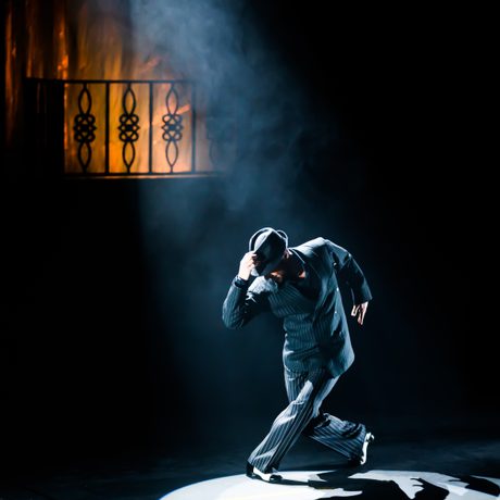 A man under a spotlight wearing a hat. He is bending down with one hand on his hat.