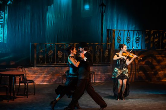 A man and a woman dancing the tango in front of a blue curtain with a lady playing the violin behind them.