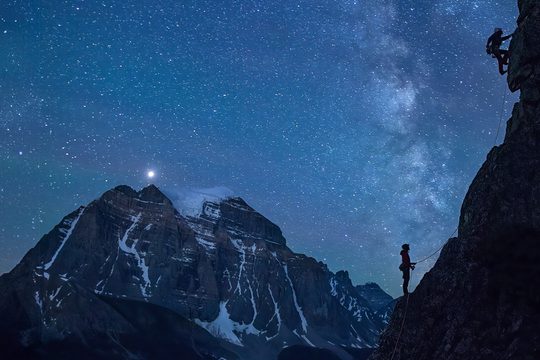 A silhouette of 2 climbers taking on a face of rock. behind them is a beautiful landscape of a mountain and the stary night sky