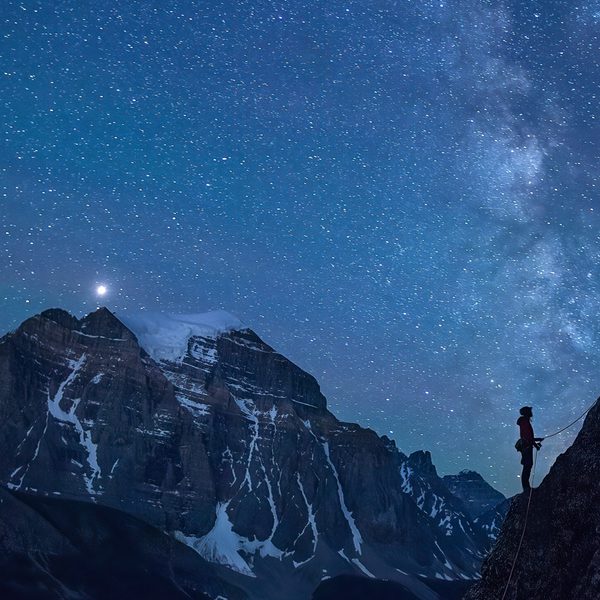A silhouette of 2 climbers taking on a face of rock. behind them is a beautiful landscape of a mountain and the stary night sky