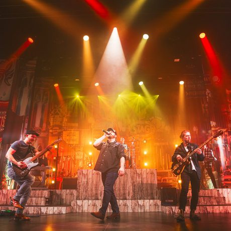 performer surrounded by two band members play on a large stage flooded with red and yellow lights from above.