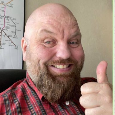 A man with a bald head and big beard, in a red and green shirt, giving a thumbs up.