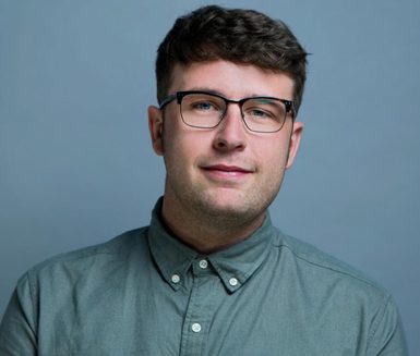 A man with brown hair and glasses, wearing a blue button-up shirt.