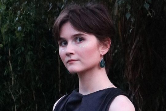A lady with a brown pixie cut standing in front of a tree, with glitter on her cheeks.
