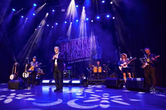 6 performers on stage playing various instruments. A white haired man stands at the front singing