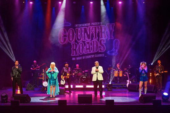 four performers stand singing to the audience, behind them the band plays