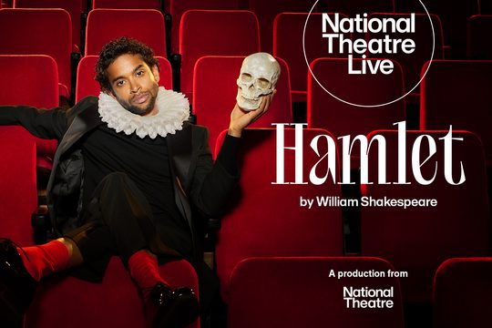 Man in a suit and tie wears an Elizabethan ruff, holding a human skull sat in red theatre seats.