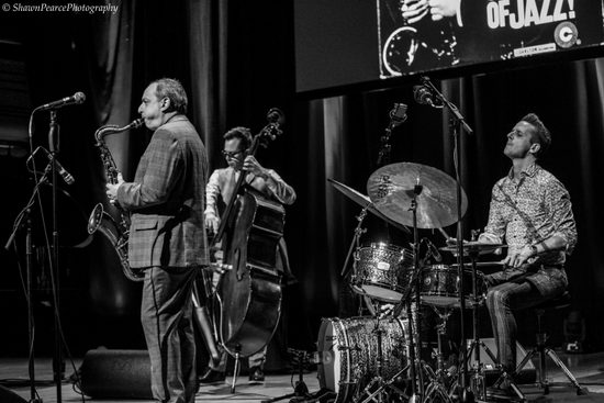 three men playing drums, double bass and saxophone in black and white