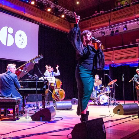 Woman singing with one hand in the air, directly behind her a man sits at a grand piano, other band members are further across the stage.