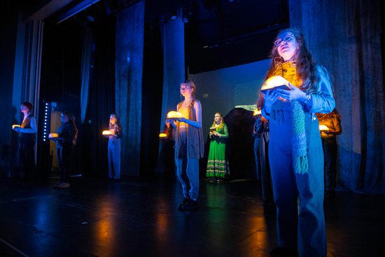 A group of people spaced out across a stage. They are all holding a book that is open and lit up.
