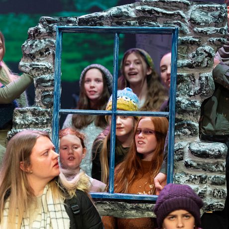 A group of people are holding up a window frame and looking through. The window frame is surrounded by grey stone.