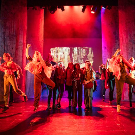 A group of dancers with masks on surround a group of women in walking clothes. The lighting is red.