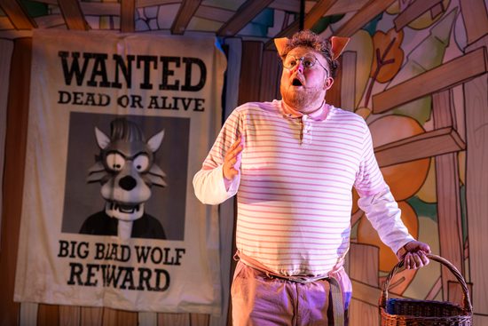One little pig in a stripy shirt is holding a basket, and looks shocked. Behind him is a wanted poster with a picture of the wolf.