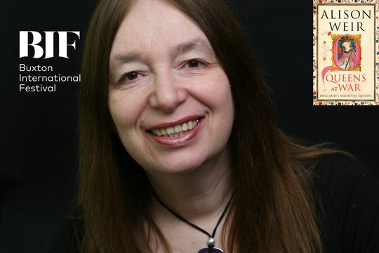 A woman with long brown hair and a large pendant necklace smiles straight at the camera.