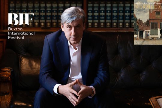 A man with grey hair in a dark blue suit sits on a leather couch with his hands together. He is in a library.