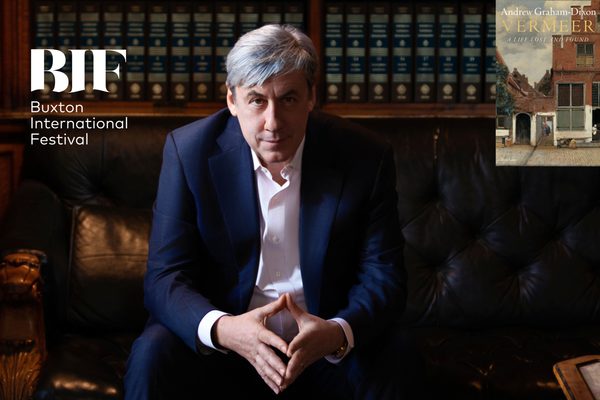 A man with grey hair in a dark blue suit sits on a leather couch with his hands together. He is in a library.