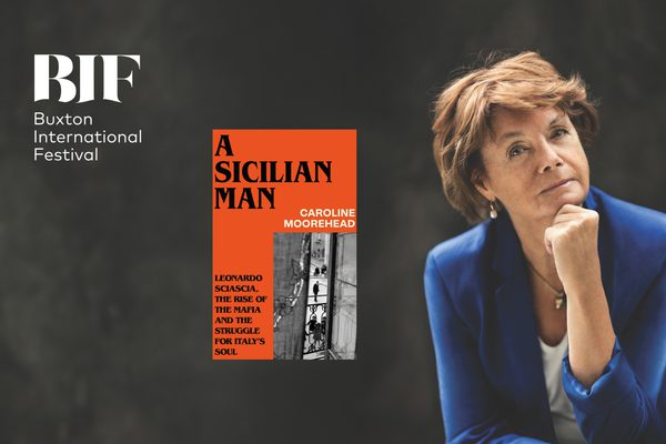 A woman with short, brunette hair is wearing a blue blazer and pearl hoop earrings with her chin resting on her knuckles against a dark, grey backdrop, a book titled 'A Sicilian Man' by Caroline Moorehead lies at the centre.